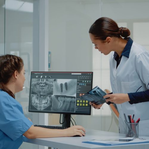 Dentist holding dentition x ray scan comparing to radiography on computer monitor at dental clinic. Nurse sitting at desk looking at screen for teeth examination and consultation